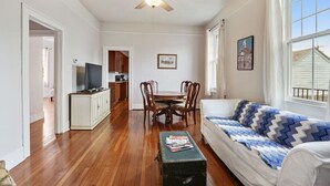 Ein Wohnzimmer mit Holzdielen, Deckenventilator, einem weißen Sofa mit gemustertem Überwurf, einem Couchtisch mit Zeitschriften und einem Essbereich mit Holzhöhlen und einem Tisch.