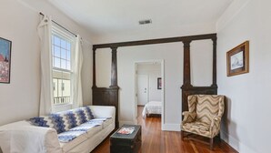 Ein Wohnzimmer mit Sofa, Sessel und einem kleinen Tisch. Ein Fenster mit Vorhängen, eine Tür zu einem anderen Raum und ein Bild an der Wand sind ebenfalls zu sehen.