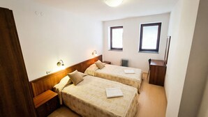 A small, clean room with two beds, a wooden nightstand, a desk, and a radiator.
