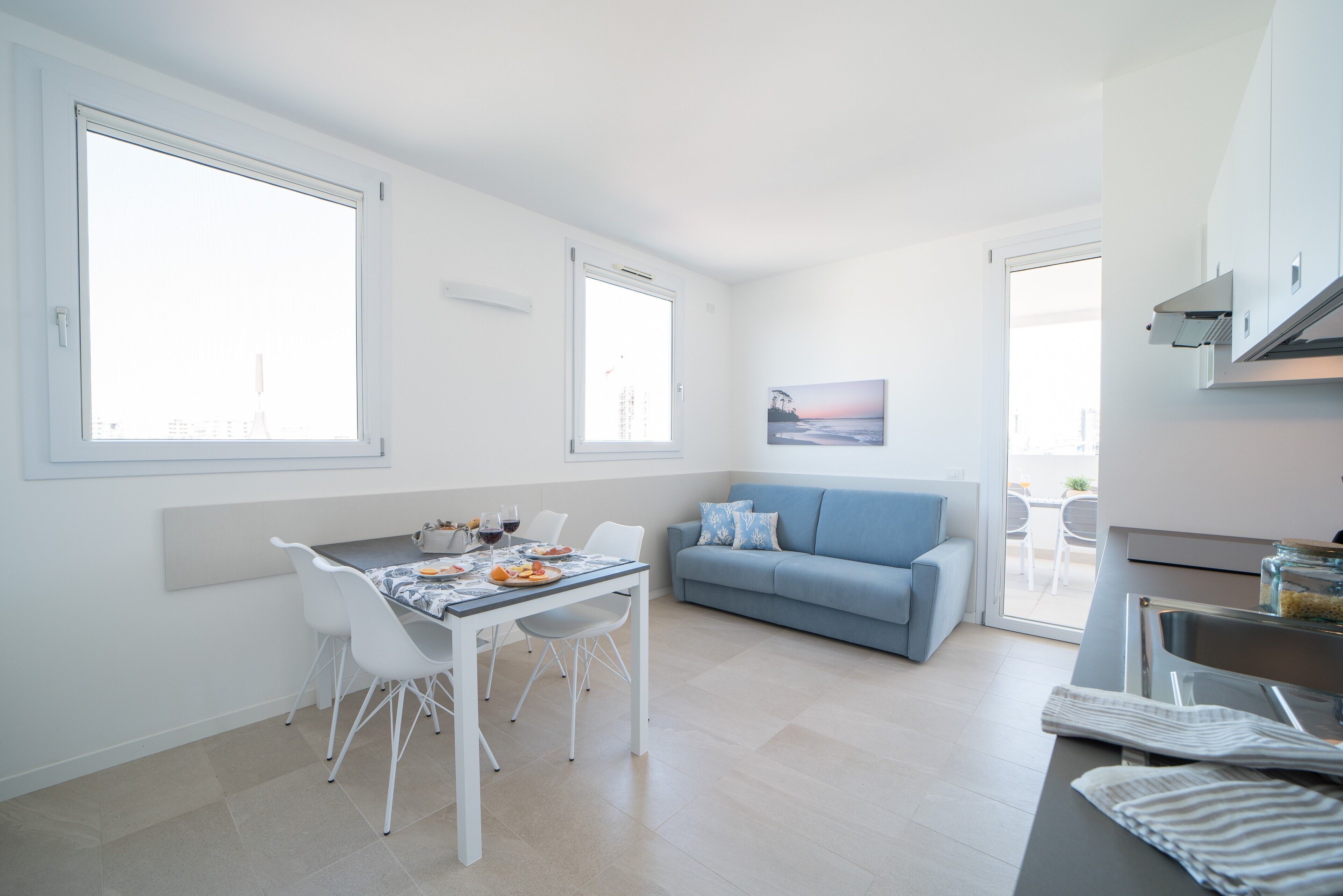 A modern kitchen with white cabinetry, a dining area with a table and chairs, a blue sofa, and a large window providing natural light.