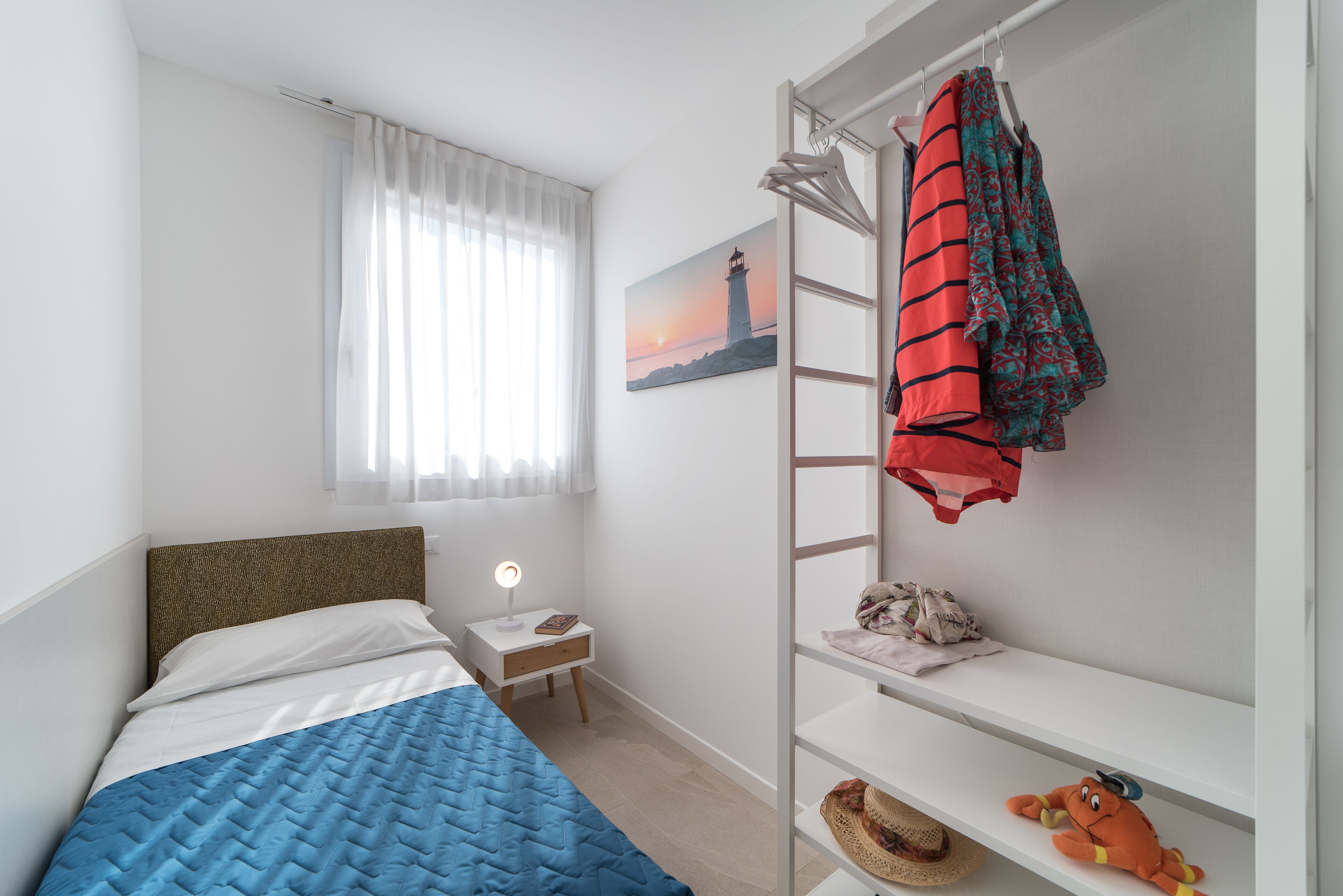 A bedroom with a bed, a nightstand, a shelf with a plush toy, and a painting of a lighthouse.