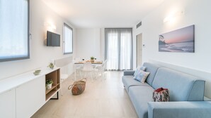 A modern living room with a sofa, a dining table with chairs, a TV mounted on the wall, and a large window with curtains.