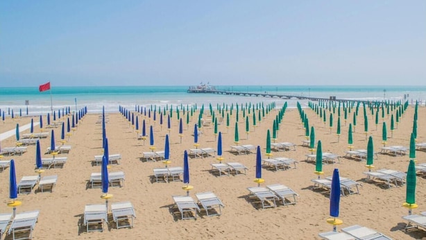 A beach with rows of sun loungers and umbrellas, a pier in the background, and a flag on the left.
