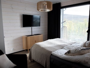 Een slaapkamer met een bed, een aan de muur bevestigde televisie, een houten kast en een raam met jaloezieën.