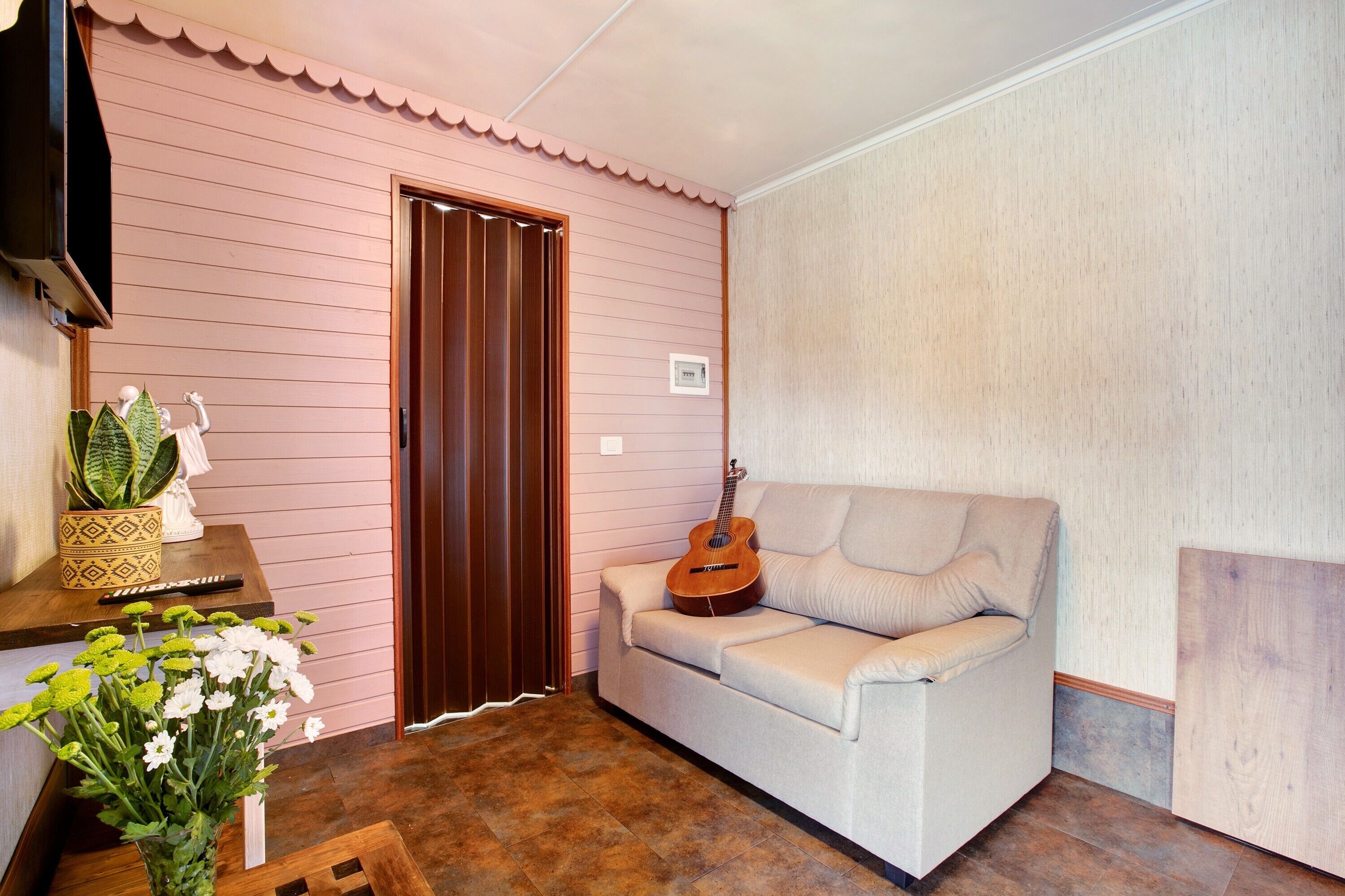 Una habitación con pared rosa, puerta de madera, sofá blanco con una guitarra, una mesita con una planta y un jarrón con flores.