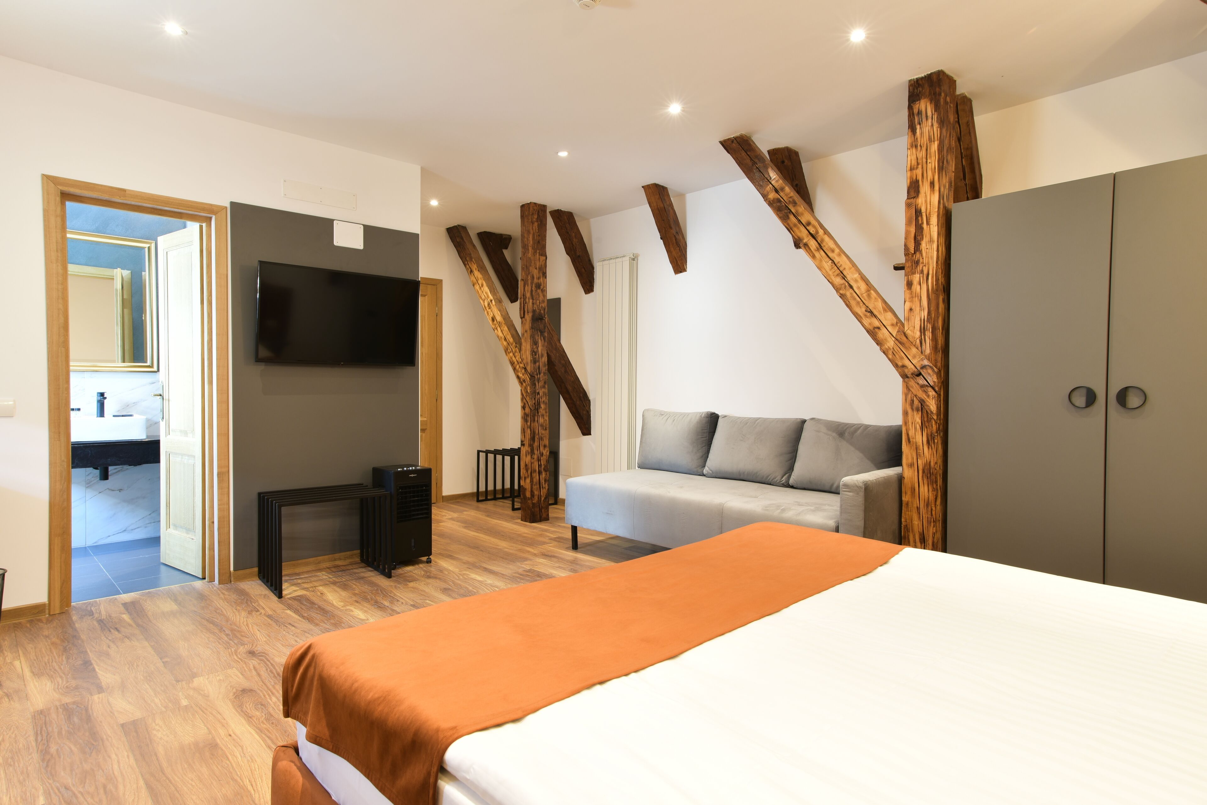 A modern bedroom with a large bed, a grey sofa, a TV mounted on the wall, and wooden beams on the ceiling.
