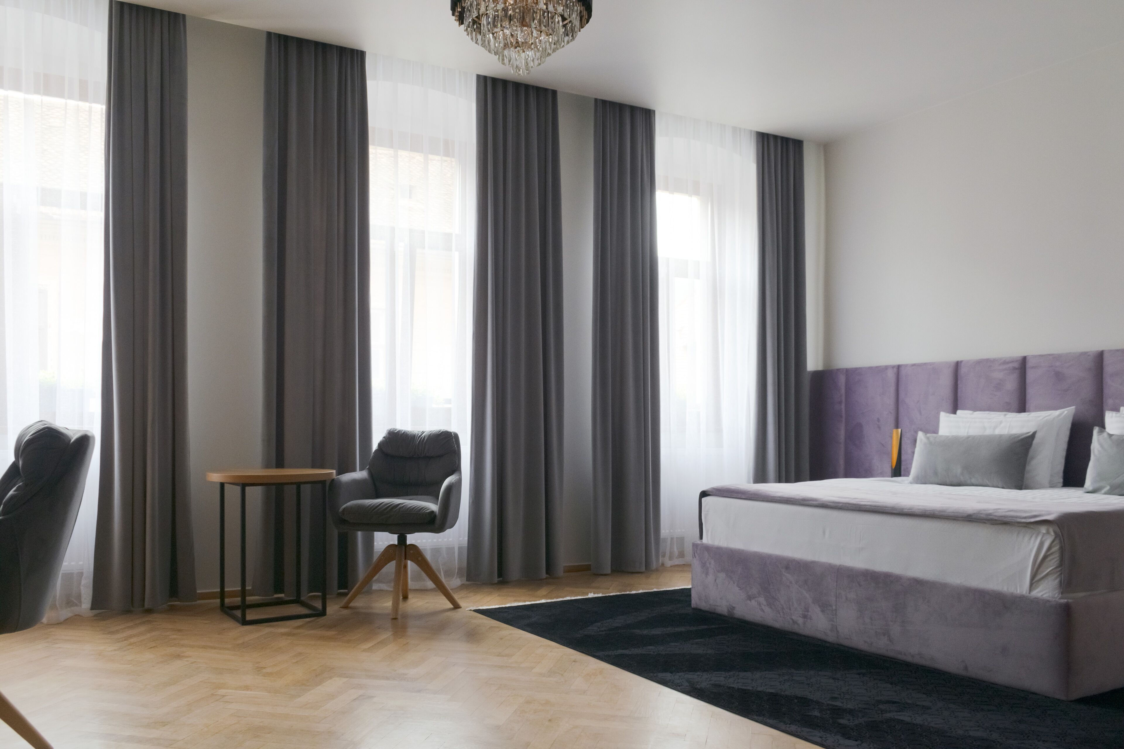A modern bedroom with a large bed, a small table, a chair, and a large window with curtains.