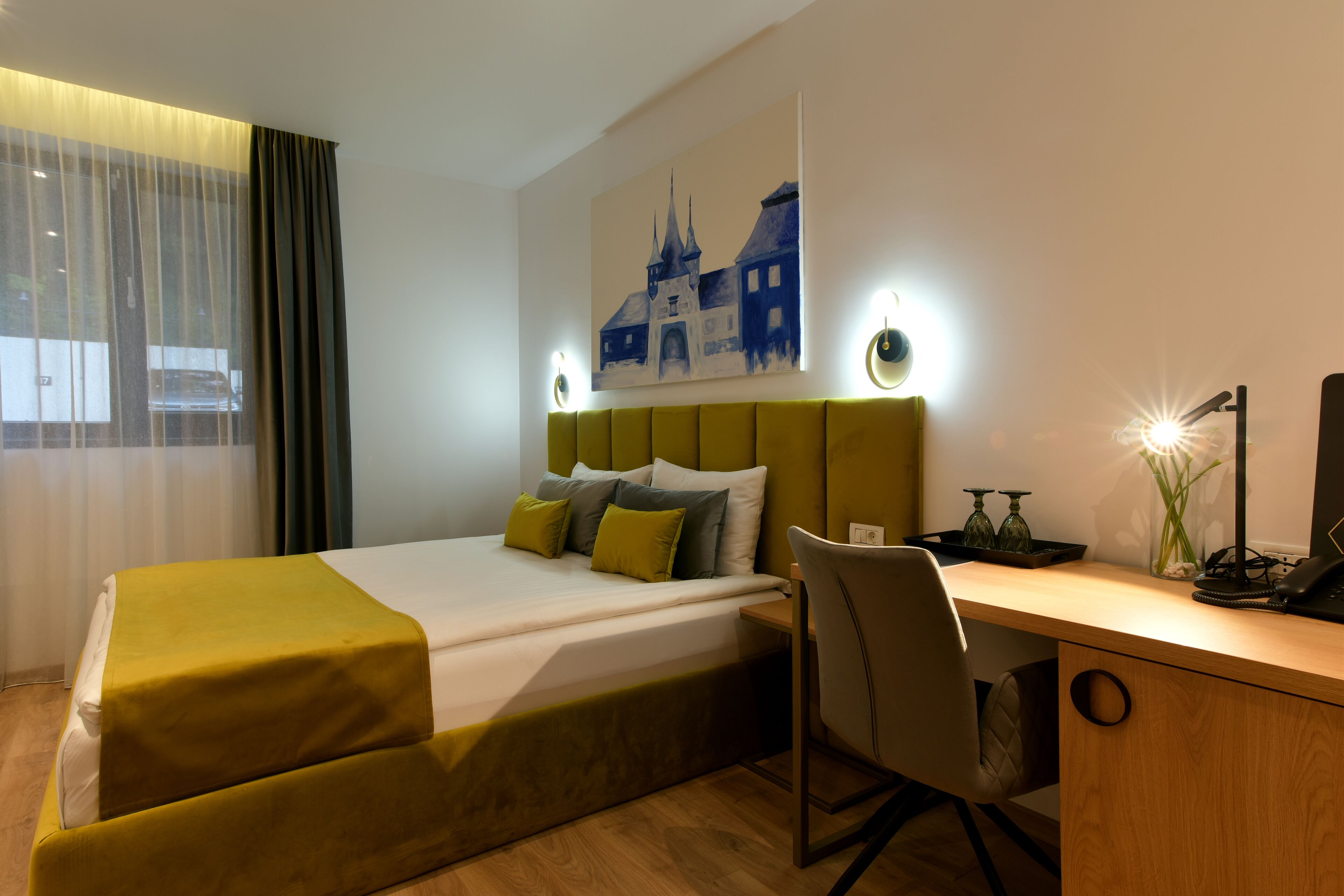 Ett hotellrum med en säng, ett skrivbord, en stol, en tavla och ett fönster med gardiner.