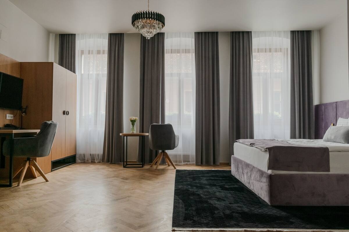 Une chambre d’hôtel moderne avec un plancher en bois, un lit doté d’une tête de lit sombre, un bureau avec une chaise, une petite table avec un vase et un lustre.