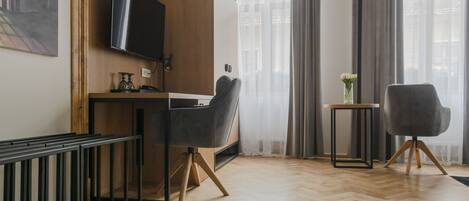 Une chambre d’hôtel moderne avec un plancher en bois, un bureau doté d’un téléviseur à écran plat, deux chaises, une petite table avec un vase et une fenêtre avec des rideaux.