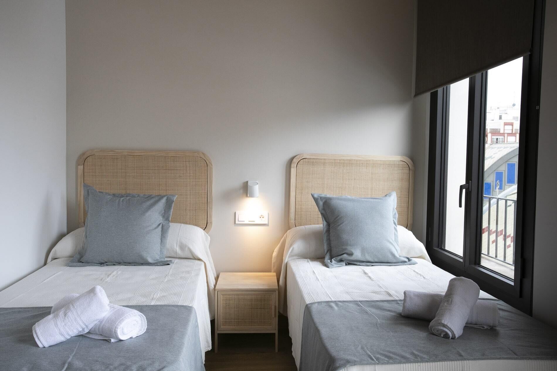 A hotel room with two beds, each with a headboard, a bedside table, and a lamp. There is a window with a view of buildings outside.