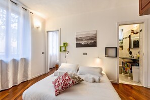 Une chambre à coucher avec un lit, des rideaux blancs, un tableau, et une salle de bain visible à travers une porte ouverte.