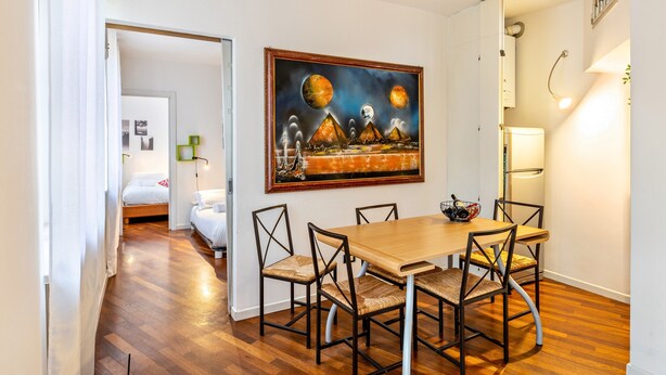 Een eethoek met een houten tafel en stoelen, een schilderij aan de muur, en een bed in de aangrenzende kamer.