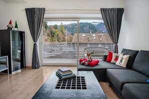 Ein Wohnzimmer mit einer Couch, einem Tisch mit Büchern, einem Schrank und Blick auf einen Balkon mit Geländer.