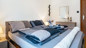 Een keurig opgemaakt bed met kussens en dekbed, een bedlampje, een dressoir met spiegel en een kapstok aan de muur.