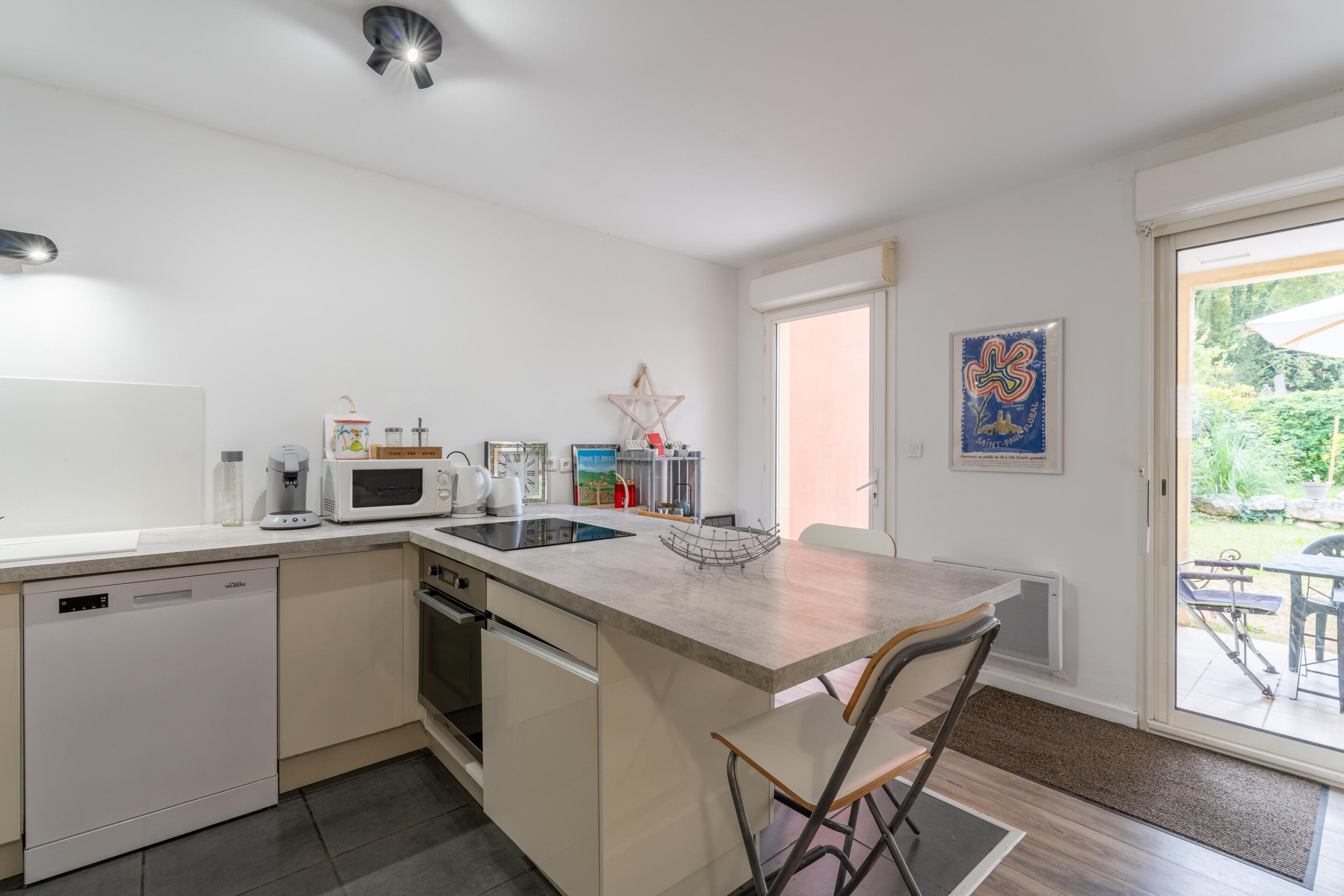 A modern kitchen with a dishwasher, microwave, kettle, and a small island with chairs.