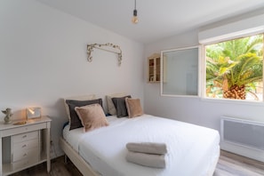 Quarto com cama, mesa de cabeceira, abajur e janela com vista para palmeiras.