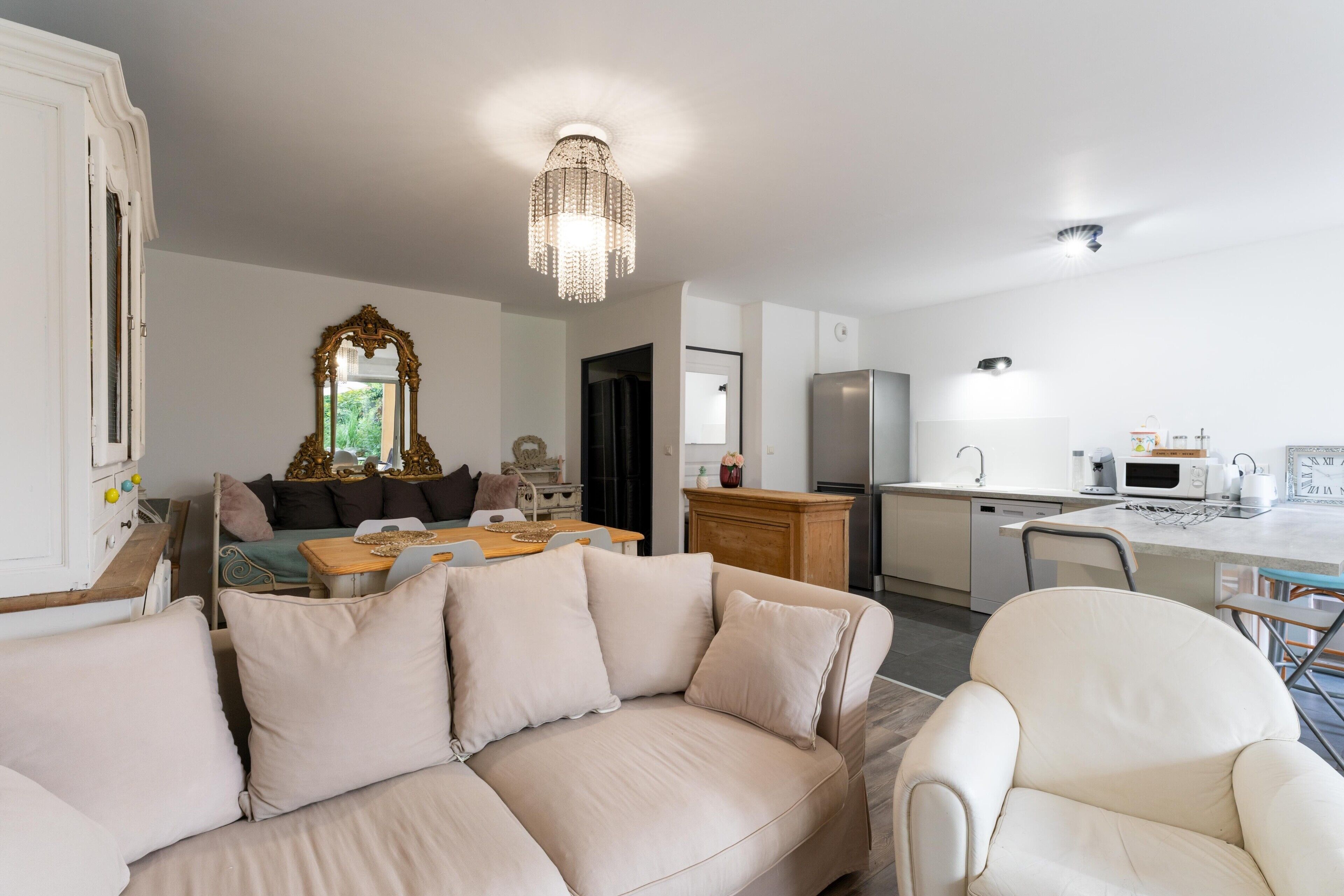 A modern living room with a beige sofa, a dining area with a wooden table, a kitchen with white appliances, and a chandelier.