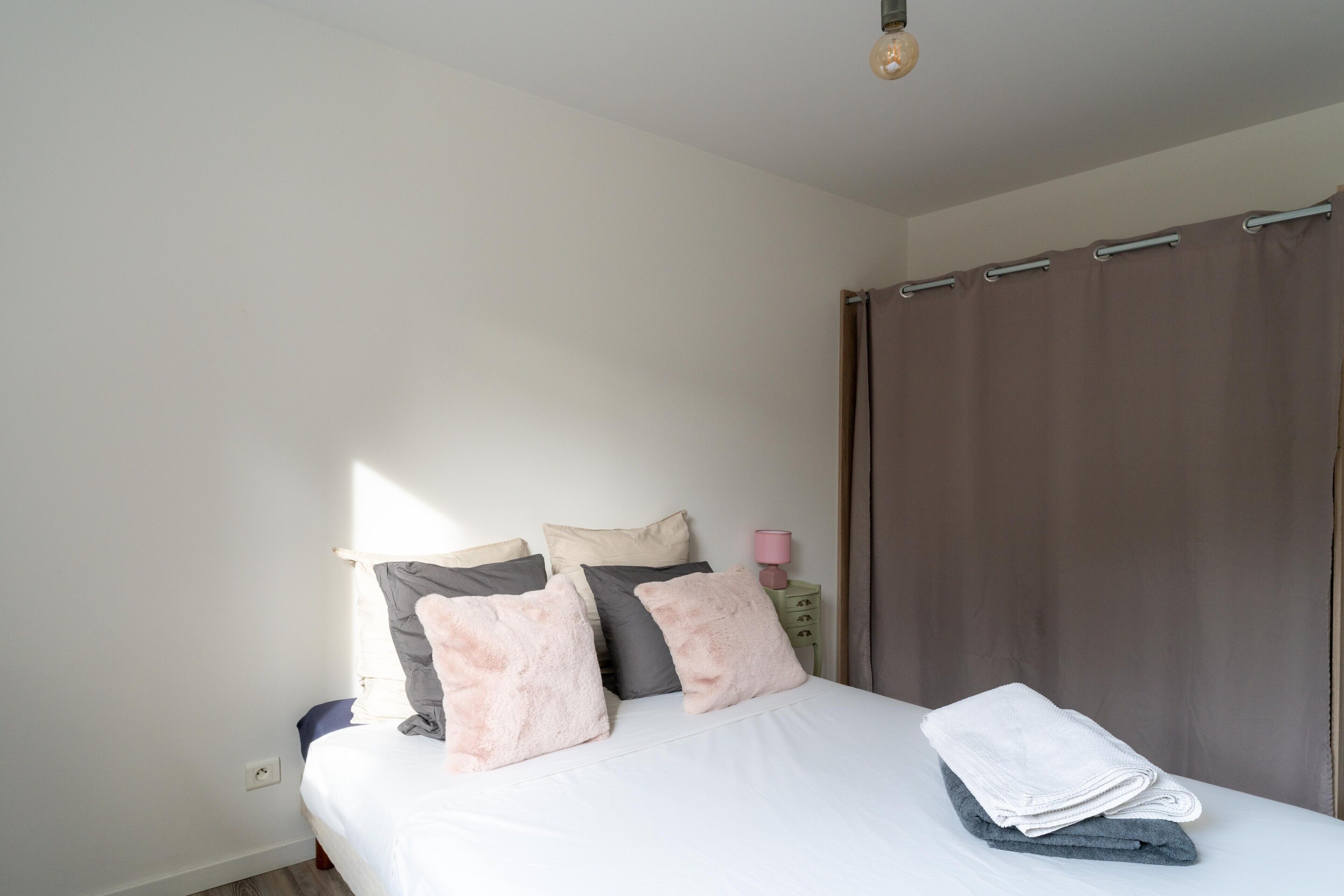 A bedroom with a bed, pillows, a nightstand, a lamp, and a curtain.