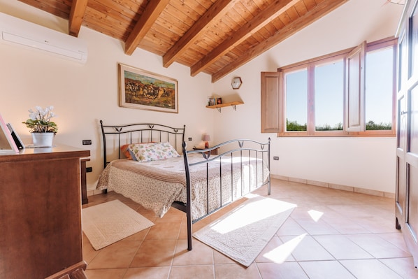 A bedroom with a bed, wooden ceiling beams, a window, a painting, and a shelf.