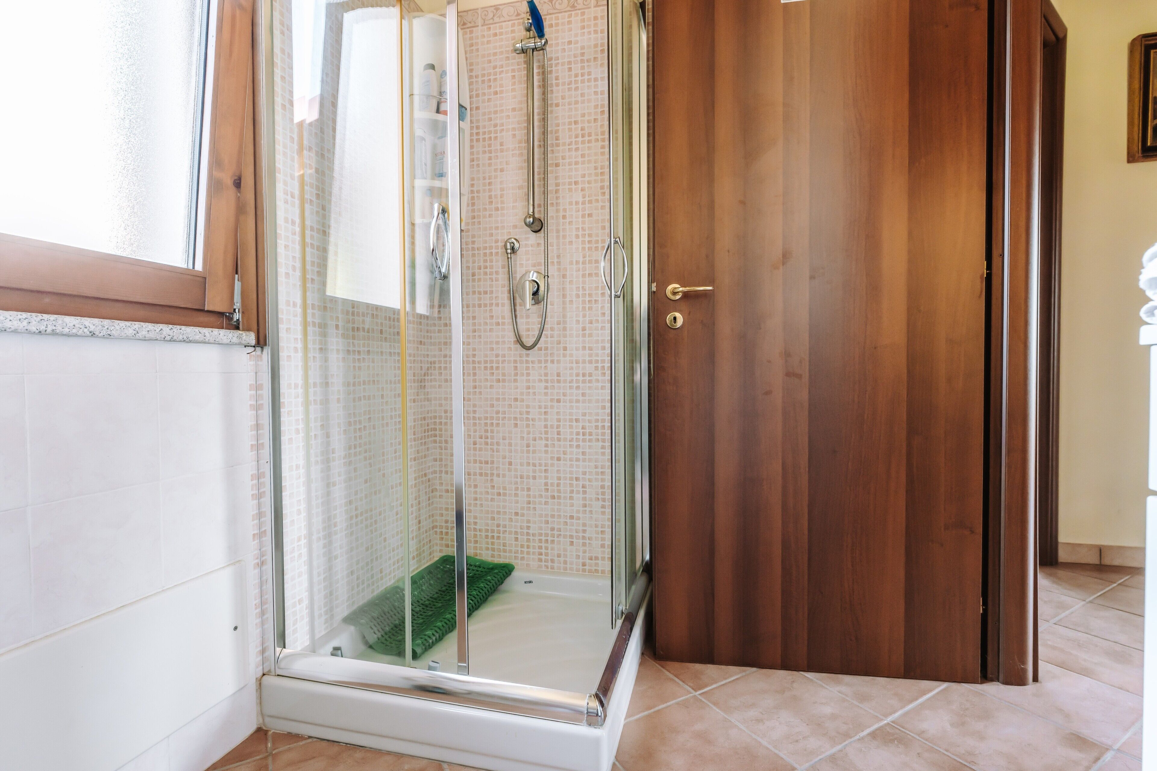 Une douche fermée par des parois en verre, dotée d’un pommeau de douche et d’un flexible de douche, d’une porte en bois et d’un sol carrelé.