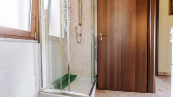 Une douche fermée par des parois en verre, dotée d’un pommeau de douche et d’un flexible de douche, d’une porte en bois et d’un sol carrelé.