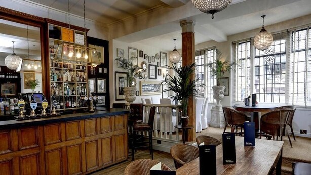 Uma área de bar com armários de madeira, cadeiras de vime e uma mesa de madeira grande com cardápios.