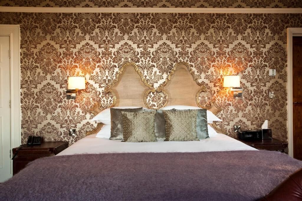 A hotel room with a large bed, ornate headboard, and two wall-mounted lamps.