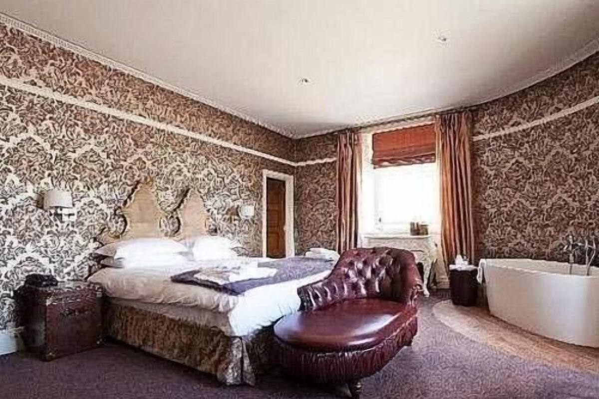 A bedroom with a large bed, a leather armchair, a bathtub, and patterned wallpaper.