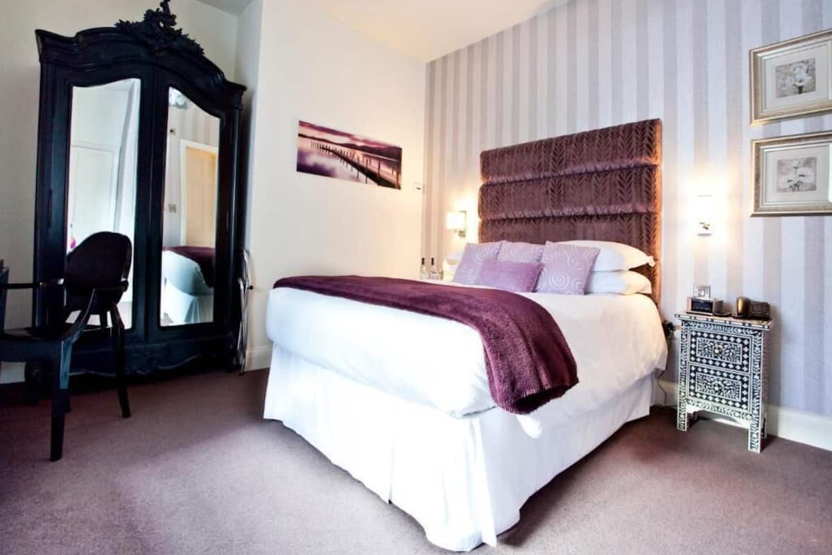 A neatly arranged bedroom with a large bed, a chair, a mirror, and a bedside table.