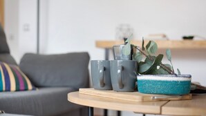 Un salon moderne avec un canapé gris, une petite table ronde en bois sur laquelle reposent deux tasses grises, et un panier en osier contenant une plante.