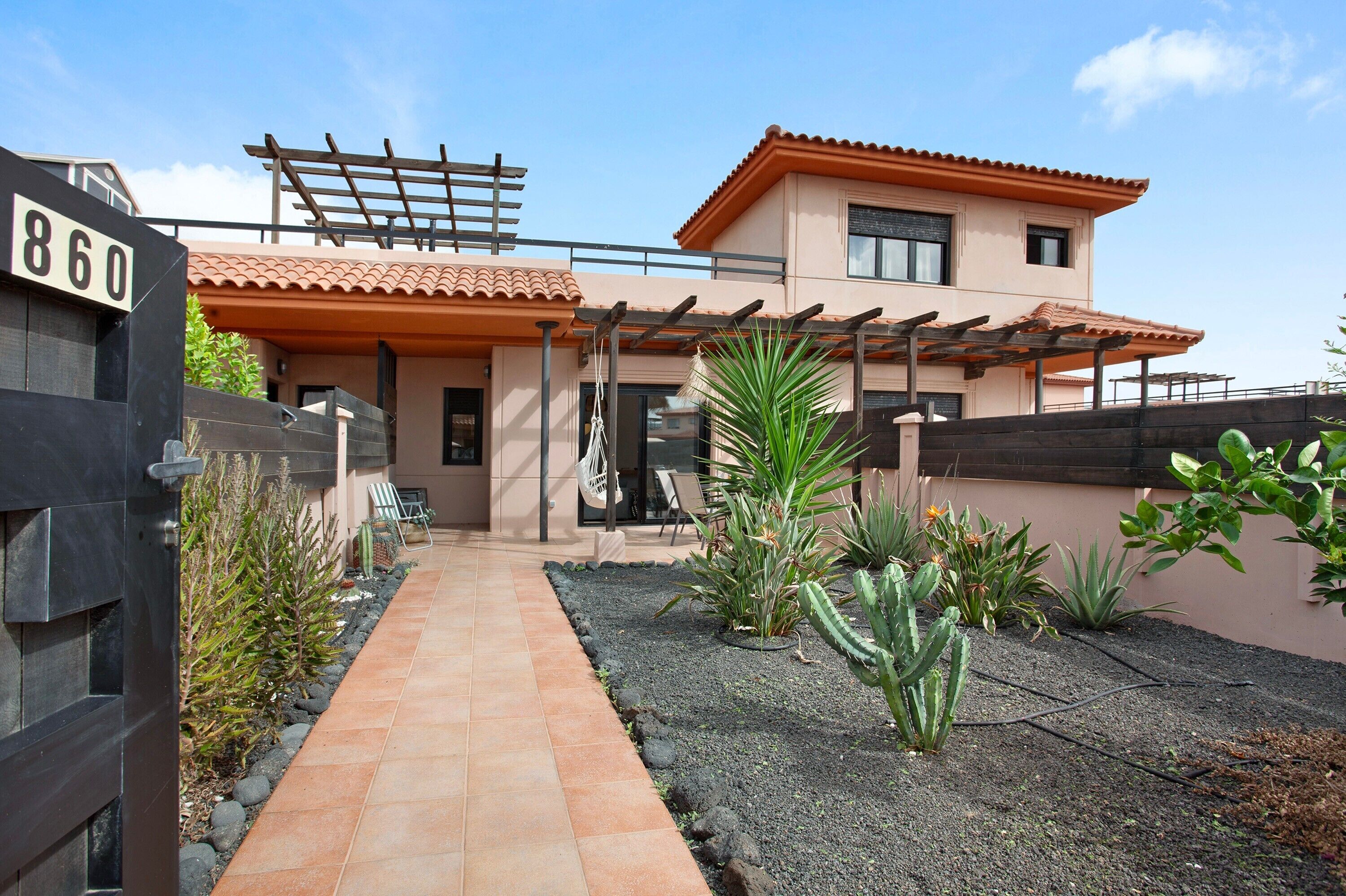 Una casa moderna de dos pisos con entrada pavimentada, un pérgola y diversas plantas.