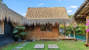 Una edificación con techo de paja, puerta y ventanas de madera, rodeada de un césped cuidado y escalones de piedra que conducen a una zona de piscina.