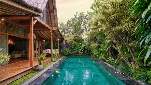 Un área junto a la piscina con cubierta de madera, un espacio de relax con sofá, y abundante vegetación alrededor.