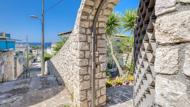 Un arco in pietra conduce a un cortile con vista sul mare, circondato da edifici e vegetazione.
