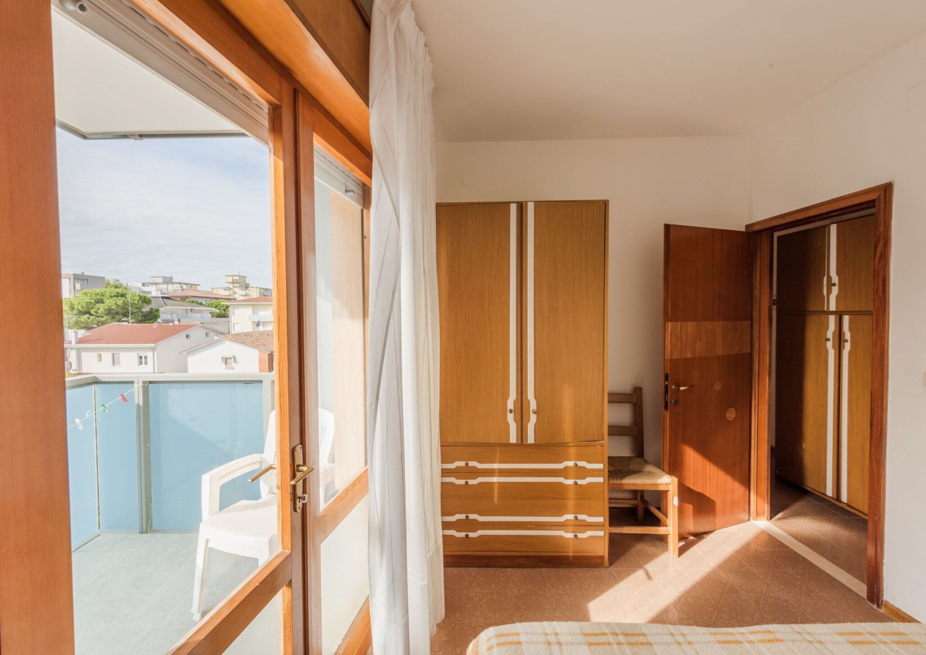Una habitación con un armario de madera, una mesita y una silla. Tiene un balcón con vistas a edificios y a una piscina.