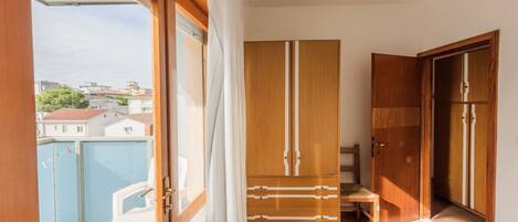 Une pièce avec une armoire en bois, une petite table et une chaise. Il y a un balcon avec vue sur les bâtiments et une piscine.