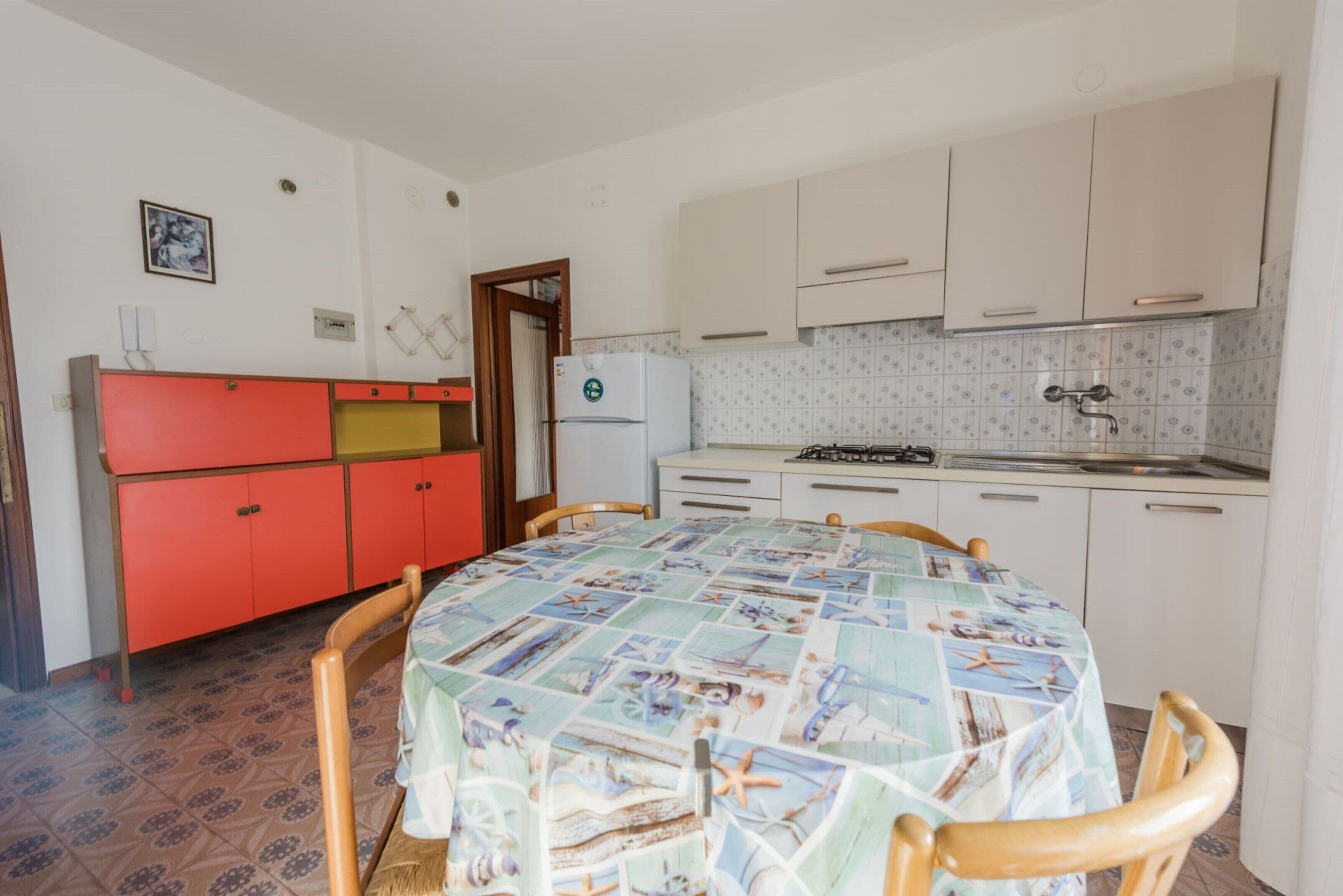 A kitchen with white cabinets, a red cabinet, a refrigerator, and a table with a nautical-themed tablecloth.