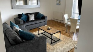 A modern living room with a grey sofa set, a coffee table, and a dining area with a glass table and white chairs. - Spacious Central Apartment (Cheshire East)