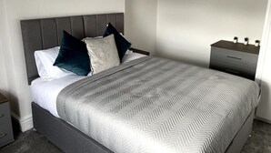 A neatly made bed with a grey herringbone patterned bedspread and pillows in white and dark blue. - Spacious Central Apartment (Cheshire East)