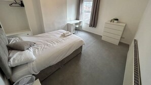 Ein Schlafzimmer mit Bett, Schreibtisch, Kommode, Heizkörper und einem Fenster mit Vorhängen.