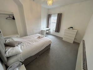 Un dormitorio con cama, escritorio, cómoda, radiador y ventana con cortinas.