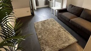 Ein Wohnzimmer mit dunklem Boden, einem braunen Sofa, einem gemusterten Teppich, einer Topfpflanze und einer Deckenleuchte.
