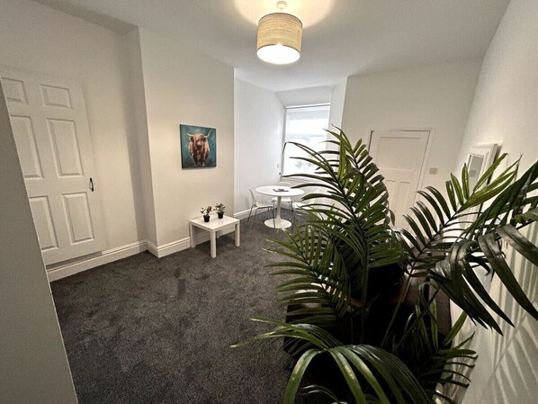 A hallway with a carpeted floor, a white door, a small table with two potted plants, a round table with chairs, and a large potted plant.