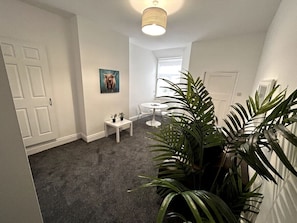 A hallway with a carpeted floor, a white door, a small table with two potted plants, a round table with chairs, and a large potted plant.