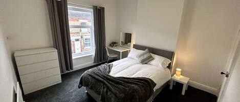 A hotel room with a bed, bedside table, lamp, desk, chair, and a window with curtains.