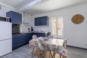 A kitchen with blue cabinets, a white refrigerator, a dining table with patterned tablecloth, and a wall clock. - Lovely vacation home Logis du roussillon (Belesta)