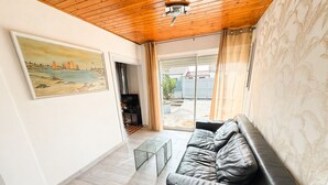 A living room with a black leather sofa, a glass coffee table, a painting on the wall, and a view through a sliding glass door. - Renovated House with Garden, Near Beach, Sleeps 4, Pet-Friendly, Private Parking (La Tranche-sur-Mer)