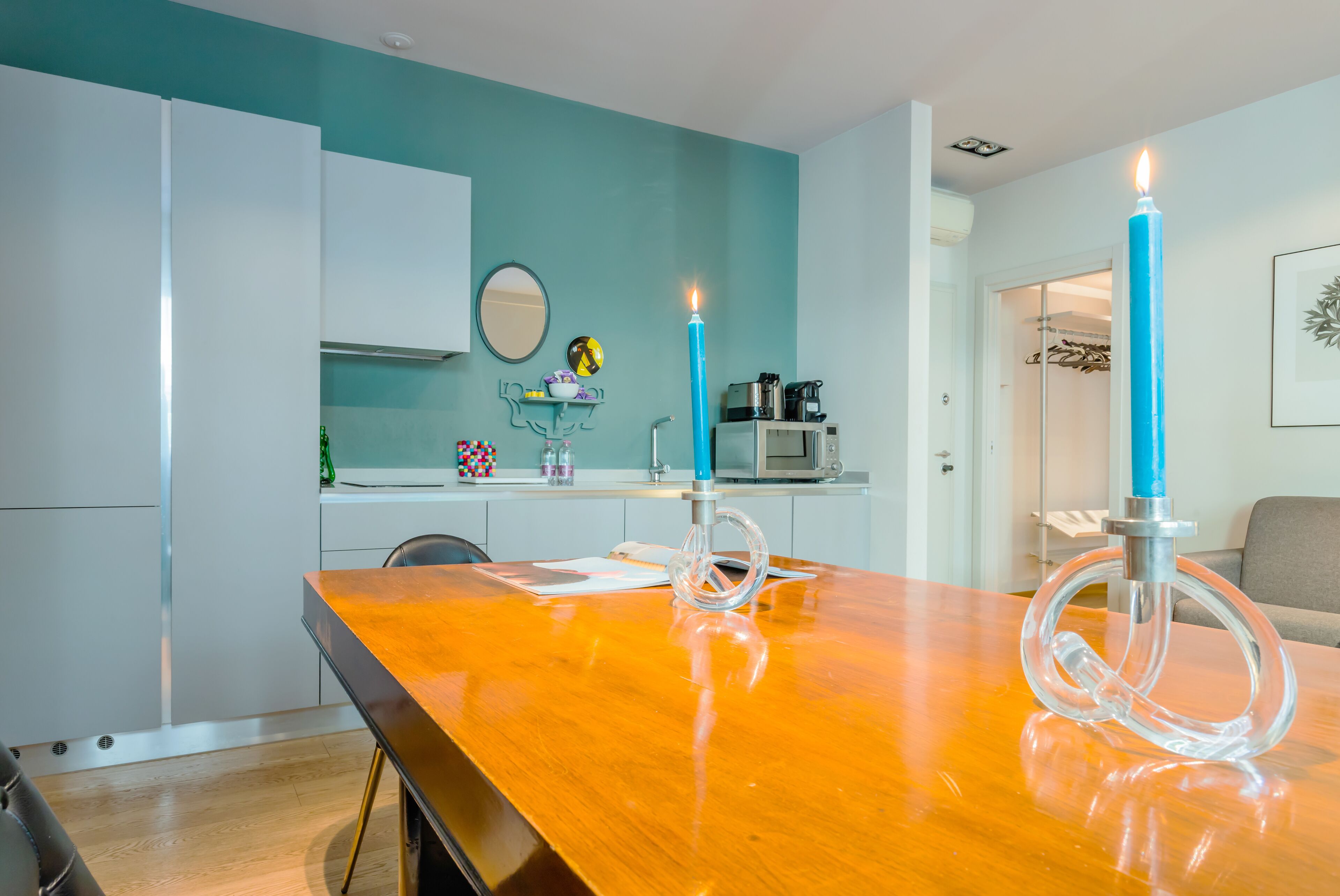 A modern kitchen with teal walls, white cabinets, and a wooden dining table with two blue candles.