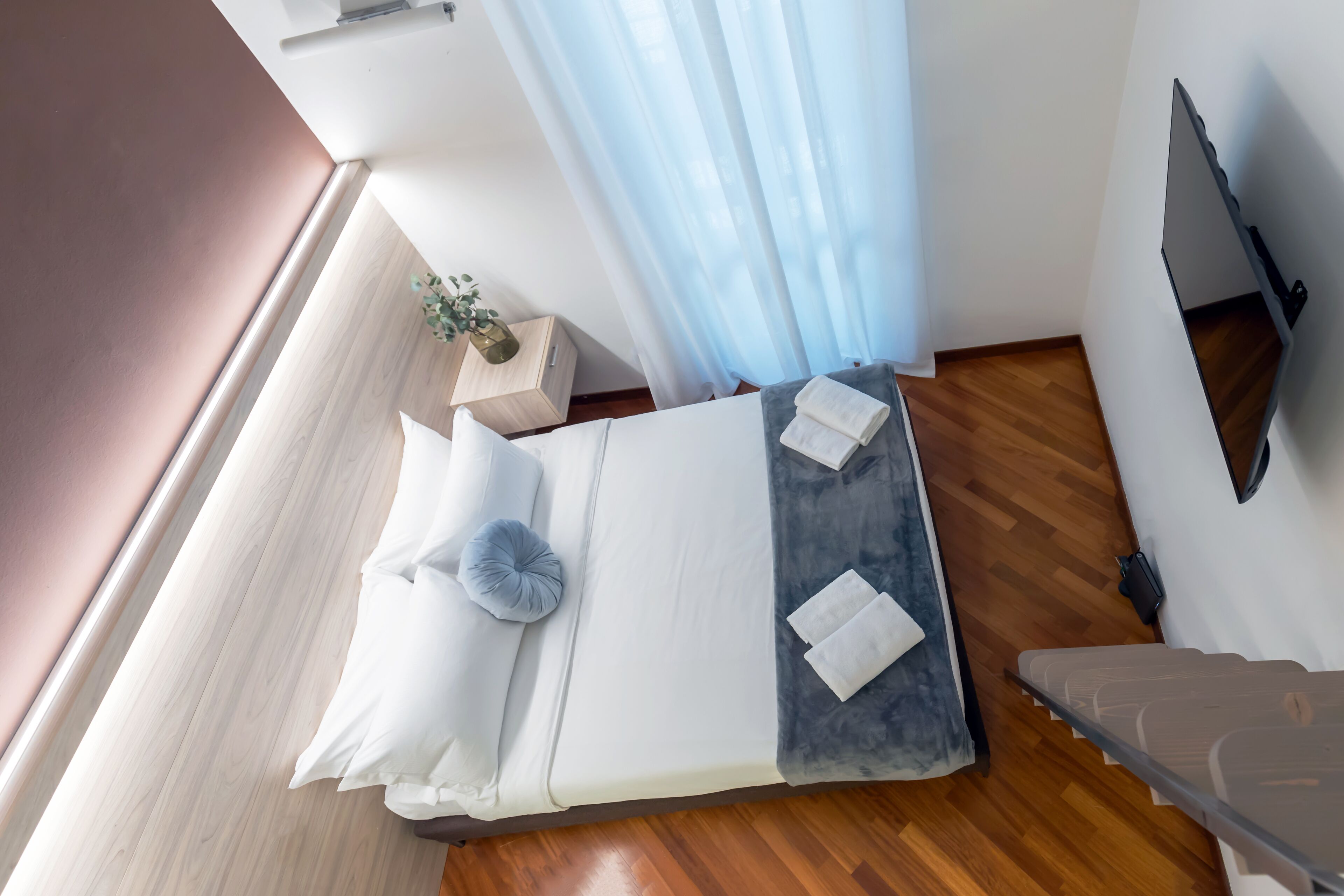 Een net opgemaakt bed met witte lakens en een grijs kussen, een klein houten nachtkastje met een plant erop, en een televisie die aan de muur is bevestigd.